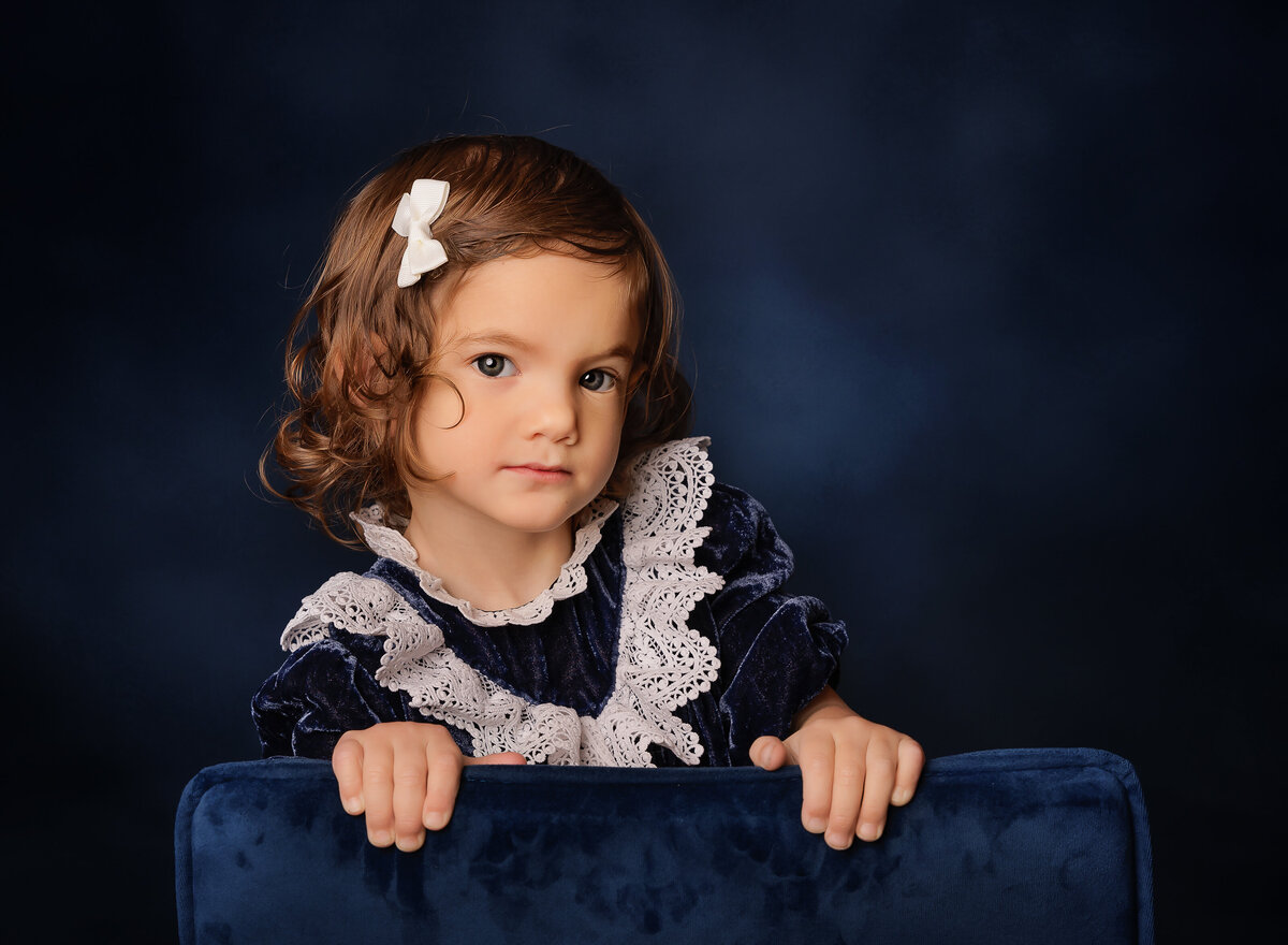 Little girl, age 2 is holding the back of a navy upholstered chair and looking at the camera with an intent gaze. Captured by Brooklyn family photographer Chaya Bornstein