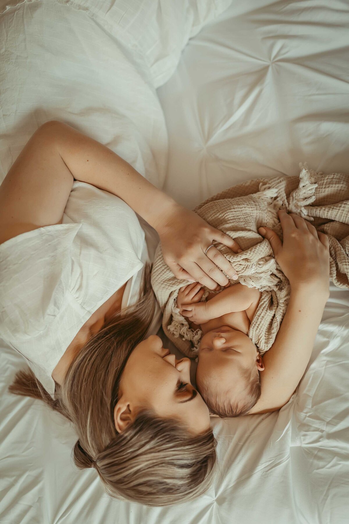 woman is laying on a bed wearing a white dress while holding her newborn baby who is sleeping and is wrapped in a light brown blanket