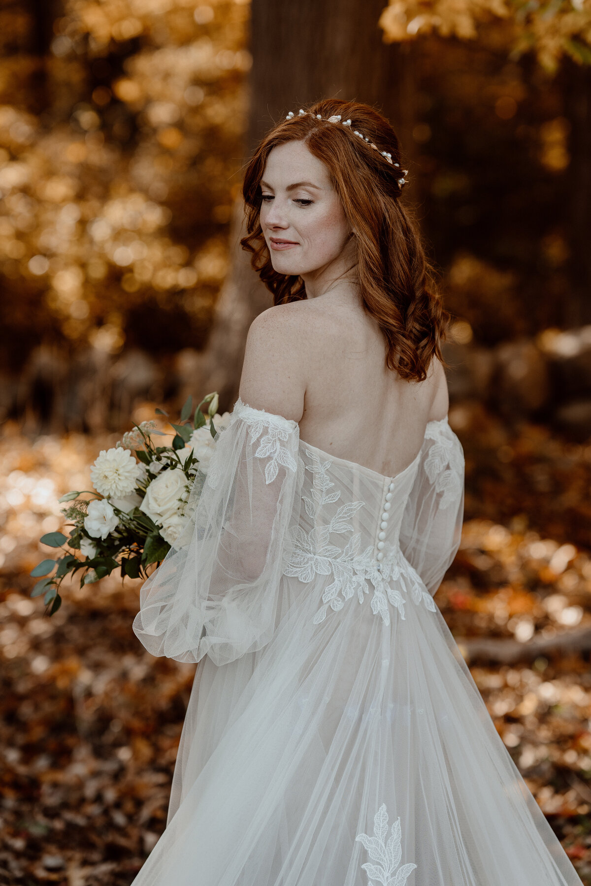 Bride with flowing red hair poses gracefully in an off-the-shoulder lace wedding gown, holding a bouquet of white flowers and greenery. Captured in a candid, documentary style, she stands amidst an autumnal forest backdrop, showcasing the natural beauty and elegance of the moment.