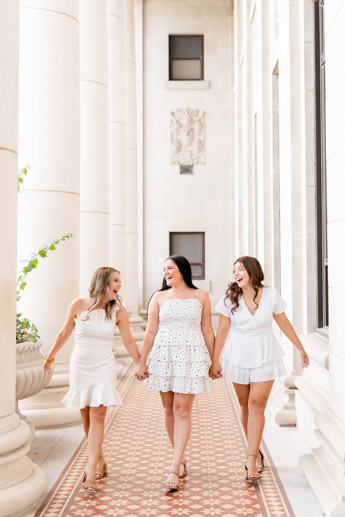 Texas A&M senior girls holding hands and walking while laughing at each other while wearing all white dresses in the columns of the Administration Building