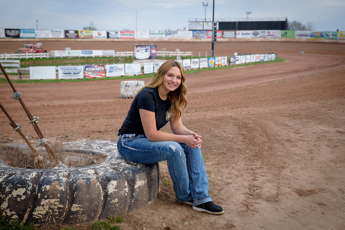 Denmark High School senior girl that loves dirt track racing.  Senior girl is sitting on tire in front of 141 Speedway race track