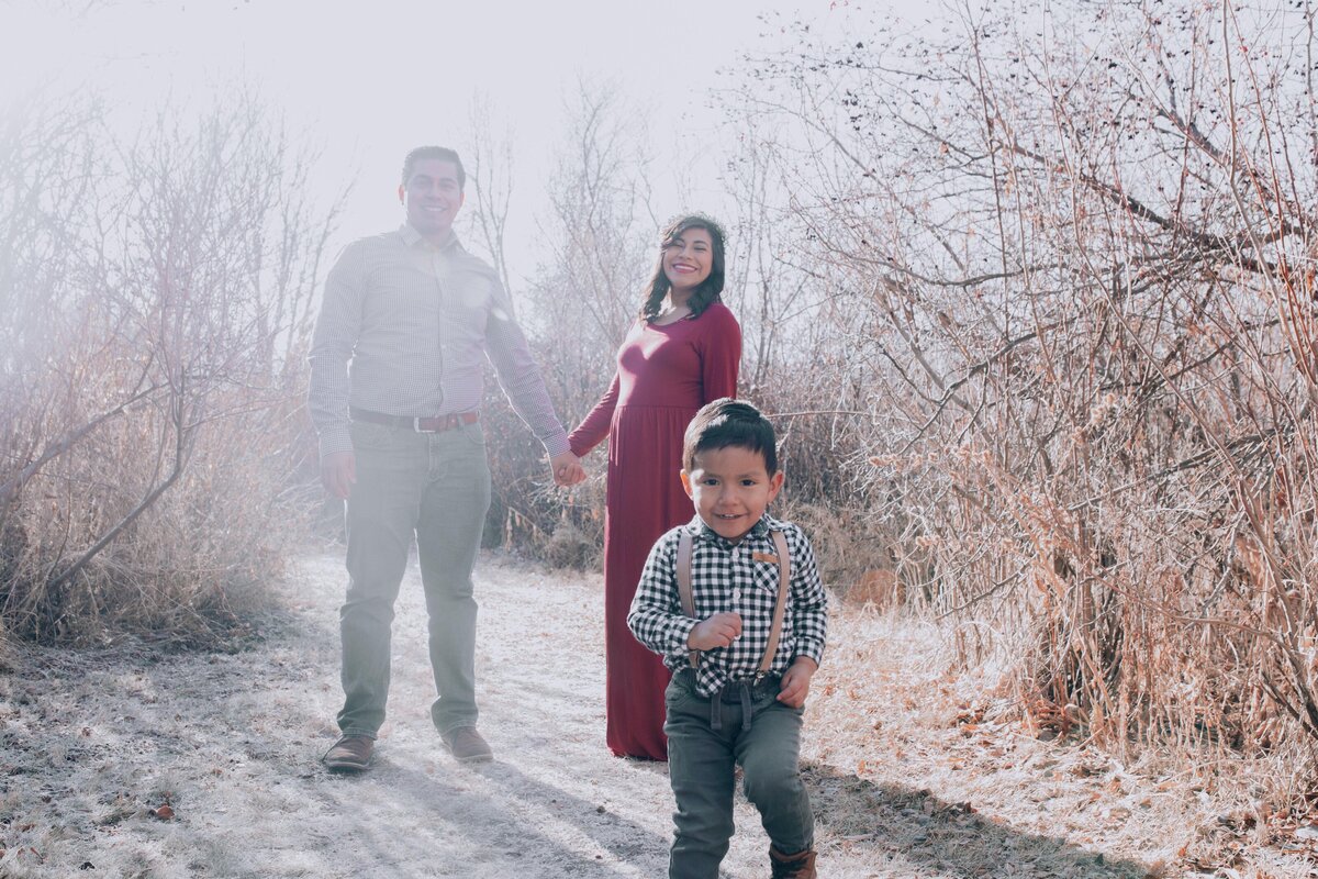 maternity photos in Idaho falls with mother and father holding hands as their toddler son runs to the camera while the sun shines in the distance as it sets behind them