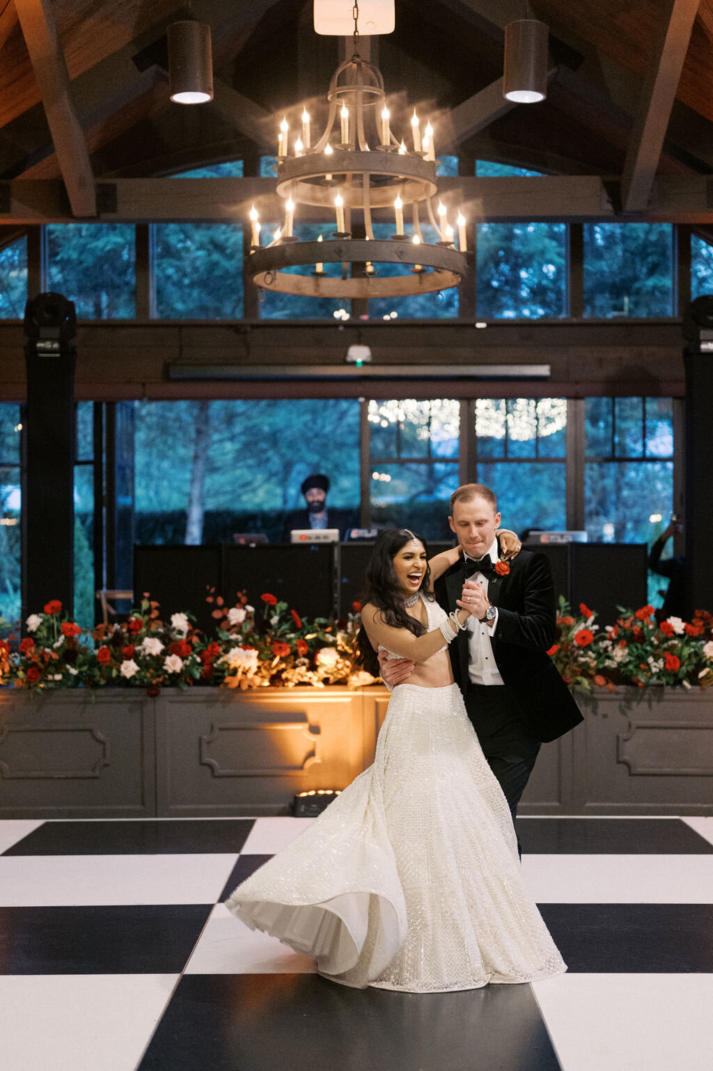 Bride and Groom share a first dance on a checkered board dance floor in front of a band stage with red florals. 