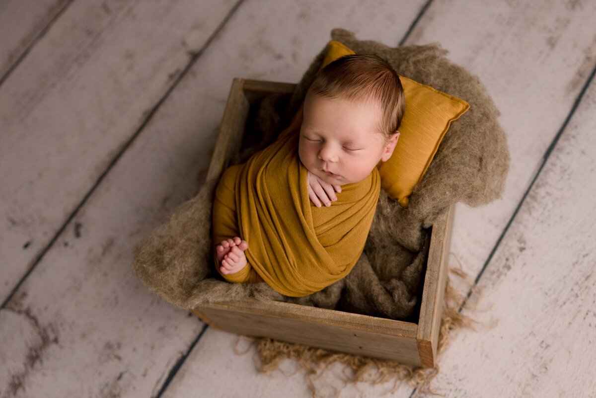 Newborn baby boy swaddled in mustard wrap and placed in a  wooden photography prop box