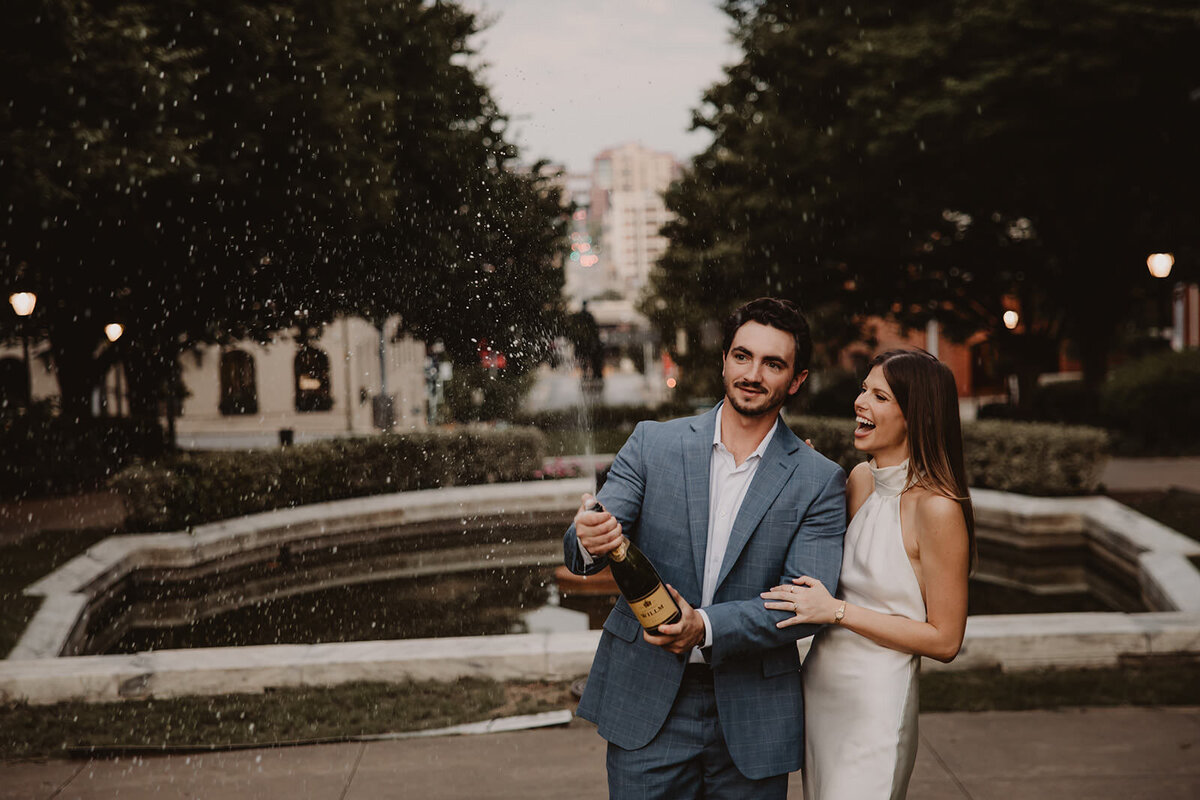 A couple standing close as one pops a bottle of champagne 