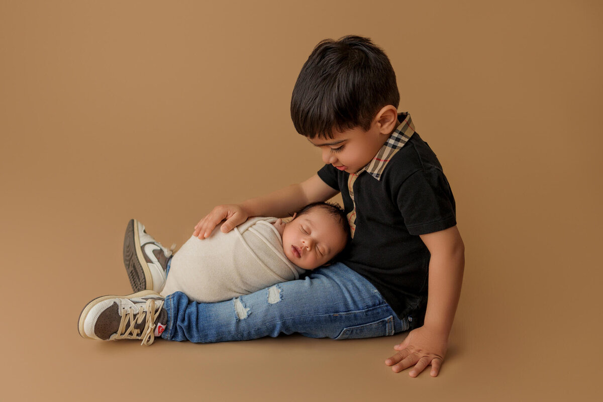 A young boy wearing a black shirt with a plaid collar and blue jeans, sitting on a tan background and gently holding a newborn wrapped in a white blanket.