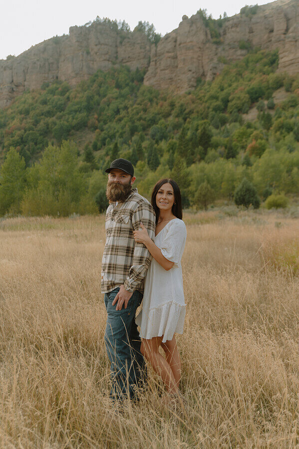 A wife holds the arm of her husband from behind as they both smile for a photo. They're standing in a tall grassy field.