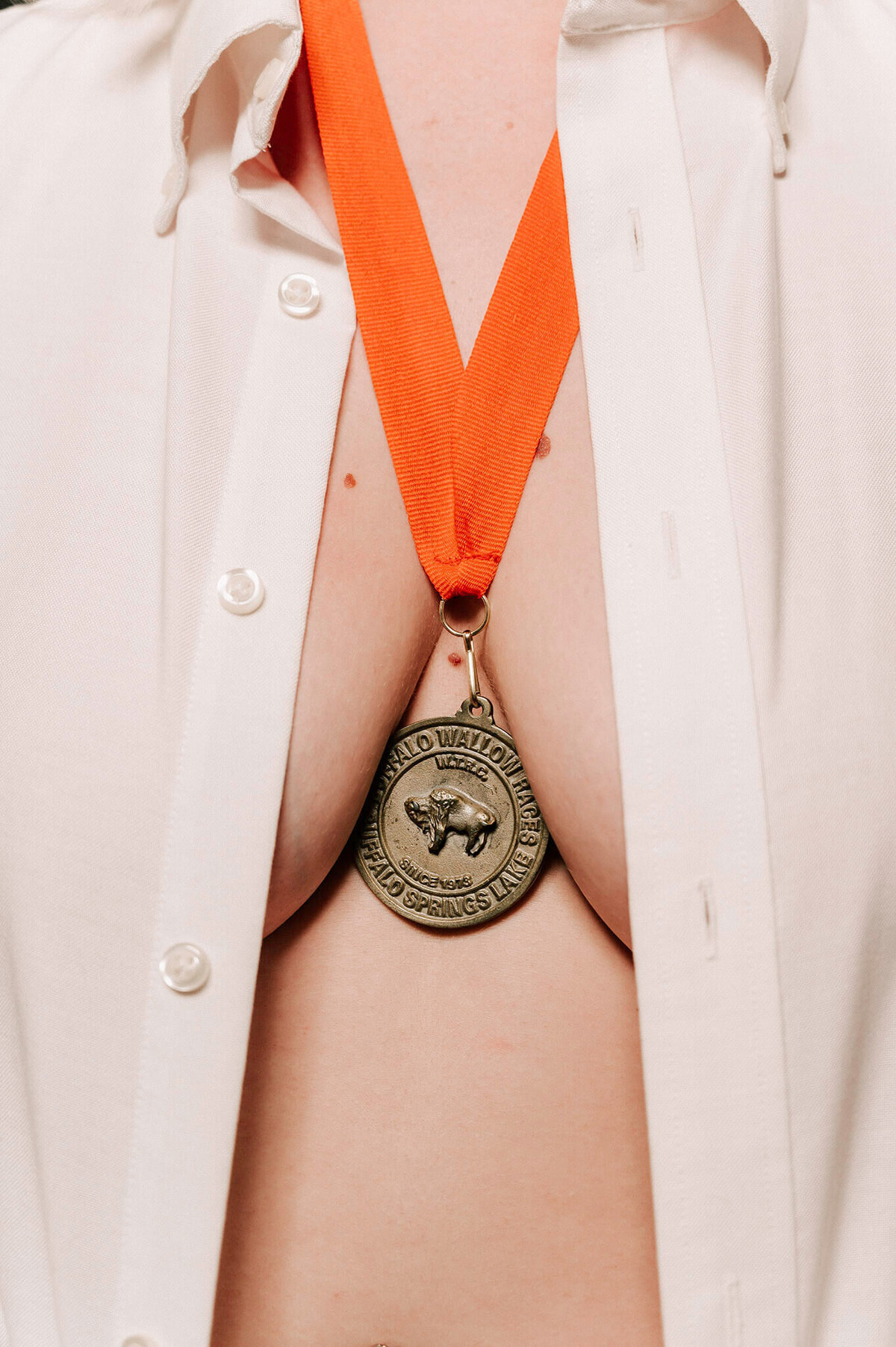Close up shot with a woman wearing a white button down shirt with it un-buttoned  and a sports metal hanging between her breasts.