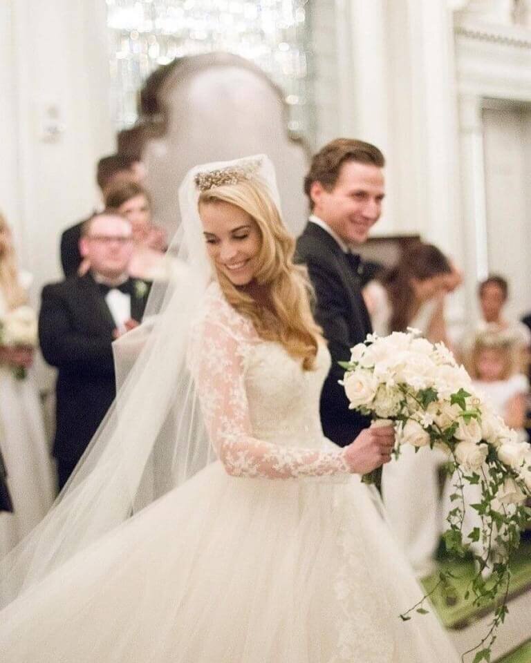 Elegant bride in a long-sleeve lace gown and cathedral veil holding a cascading white rose bouquet during a formal indoor ceremony. Mistique Makeup provides timeless bridal hair and makeup services for traditional weddings in Austin, San Antonio, and Dallas.