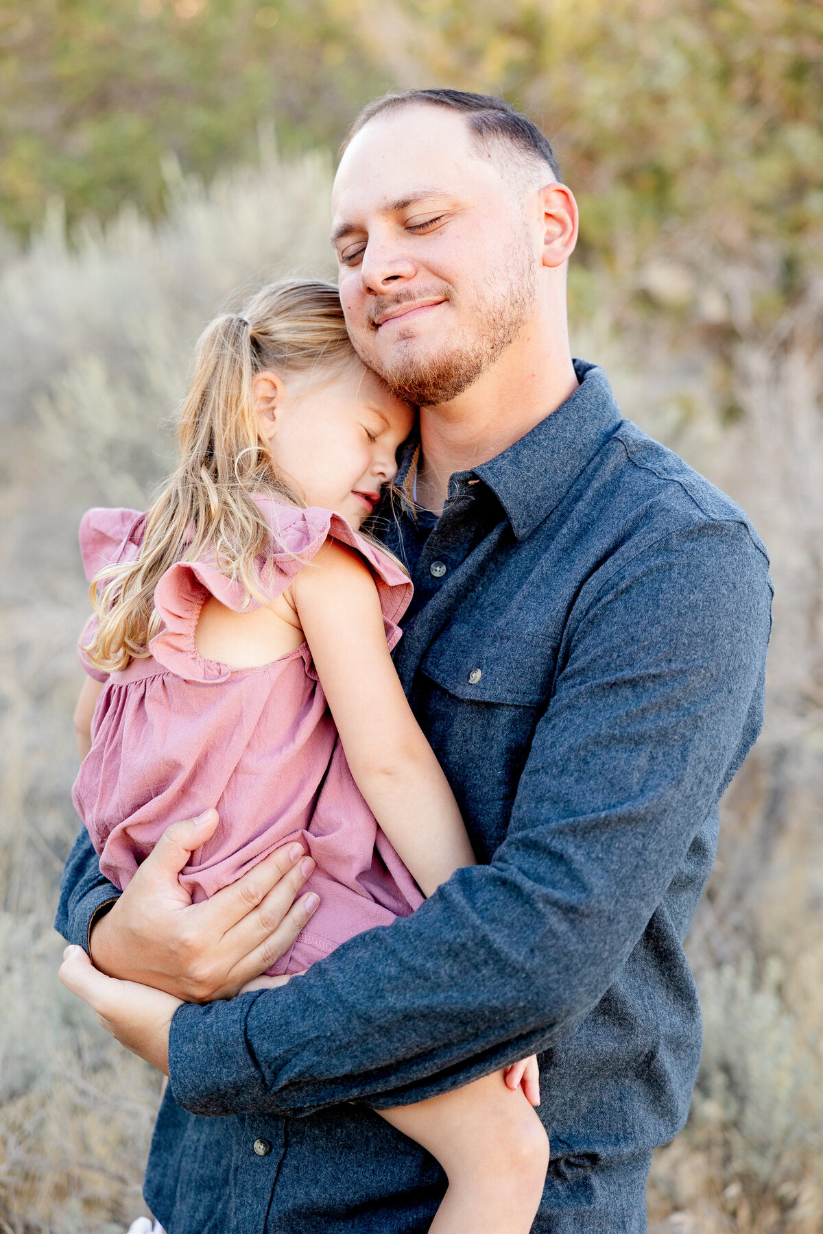 A father in a dark button-up shirt holds his daughter close as she rests her head on his shoulder. A peaceful and tender moment captured in a natural outdoor setting.