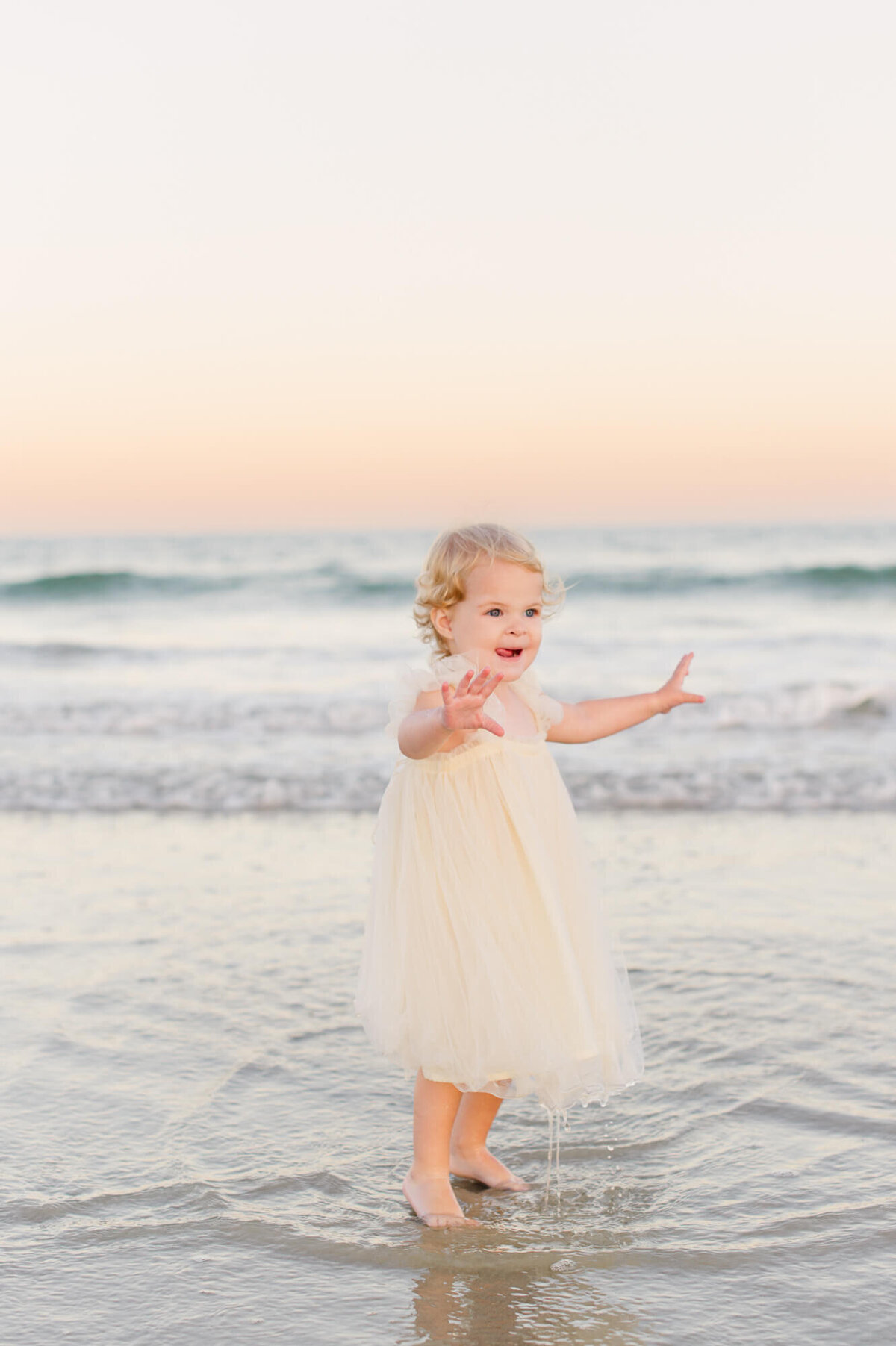 Young toddler girl plays in the ocean waves at the end of their maternity photo session