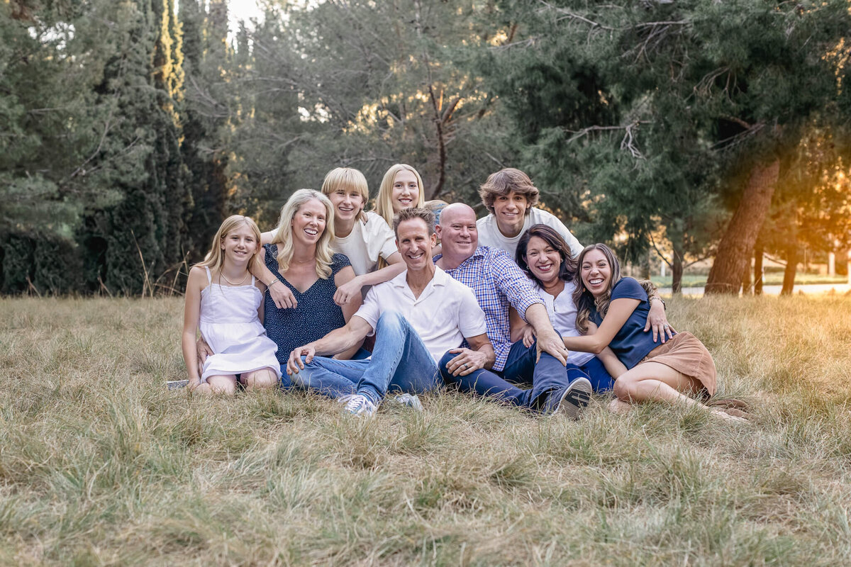 A big family sits in the grass at Jeffrey Open Space, laughing, smiling, and holding one another with green trees behind them.