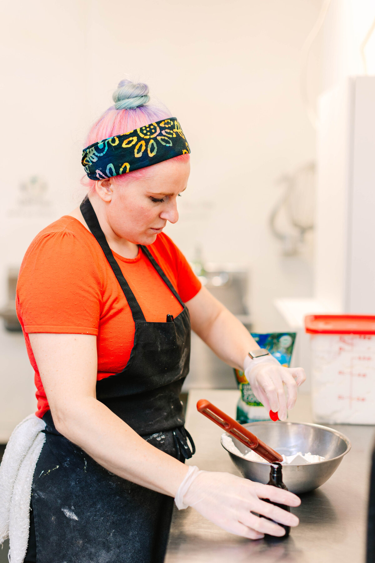 A baker with pastel-colored hair, wearing a vibrant headband, orange shirt, and black apron, carefully measures an ingredient into a mixing bowl in a commercial kitchen. She wears gloves and a smartwatch while working.