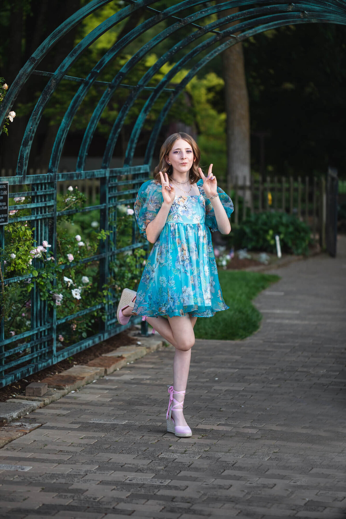 Fun portrait of a teenage girl in a blue flora dress doing a fun pose on one leg and peace signs in a garden. Captured by Springfield, MO teen photographer Dynae Levingston.