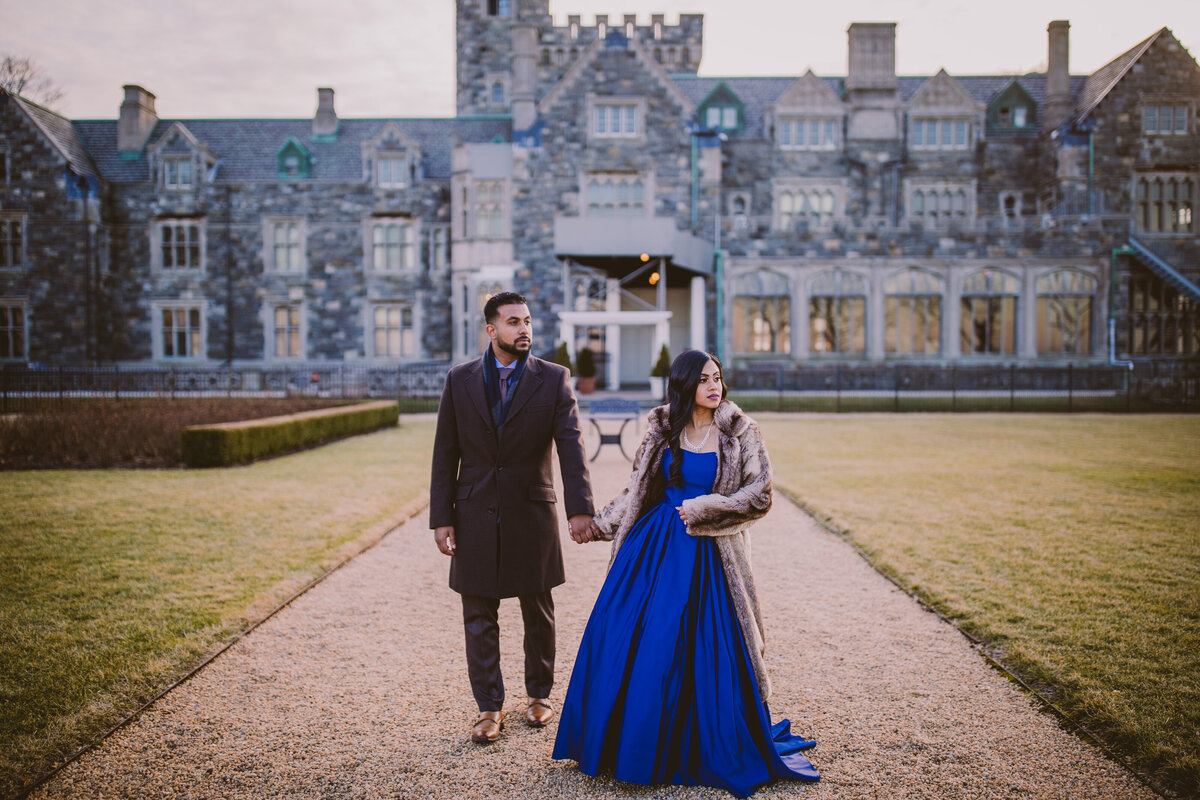 An engaged couple in formal clothing and coats walking in front of the Hempstead House mansion at Sands Point Preserve in NY