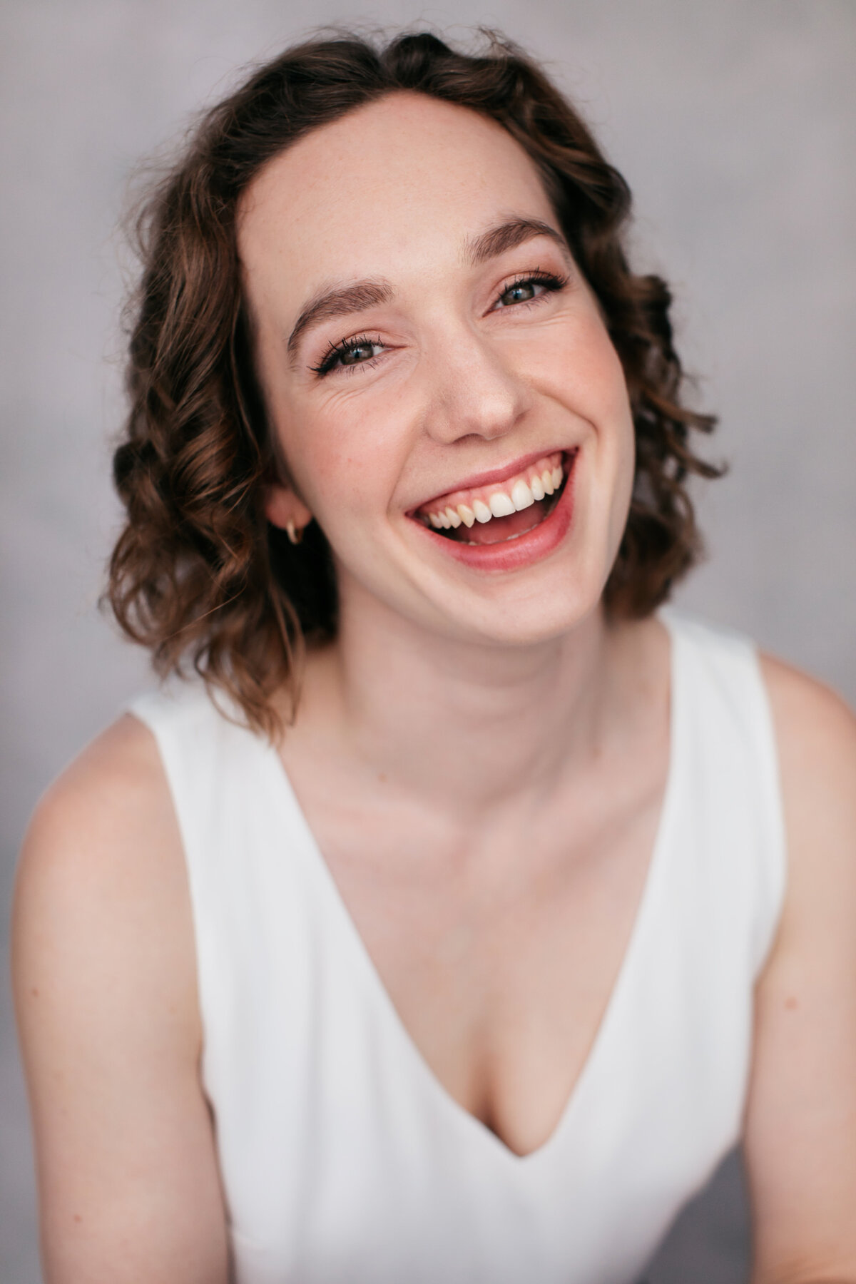Castingfoto van Emily White. Lachende vrouw met bruine krullen in een witte top voor een grijze backdrop.