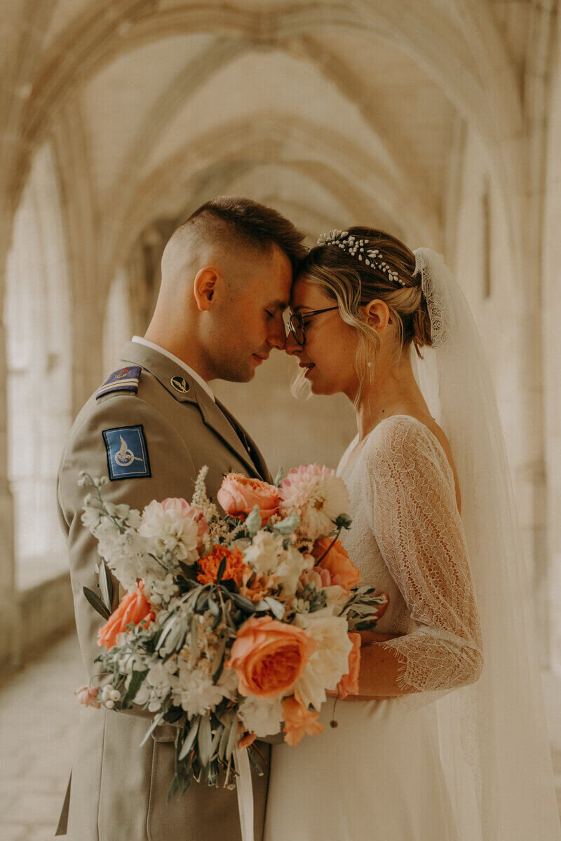 Marié en uniforme front posé sur celui de la mariée portant son bouquet, posant sous des voûtes en pierre pour Laura, photographe professionnelle en Vendée.