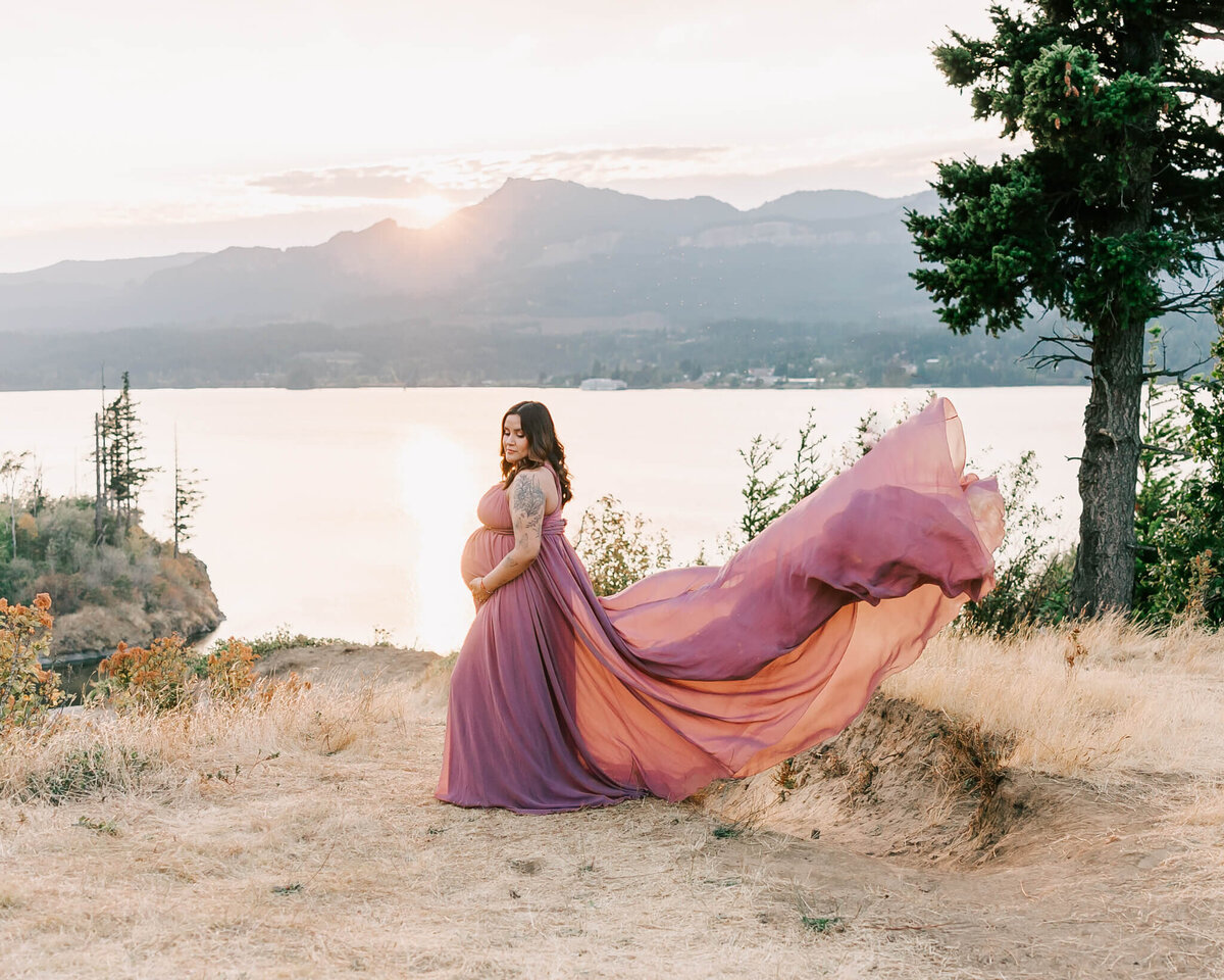 Portrait of a pregnant woman in the columbia river gorge in Oregon. She has a long pink dress and the train is flowing in the wind. The sun is setting over the hills in the distance.