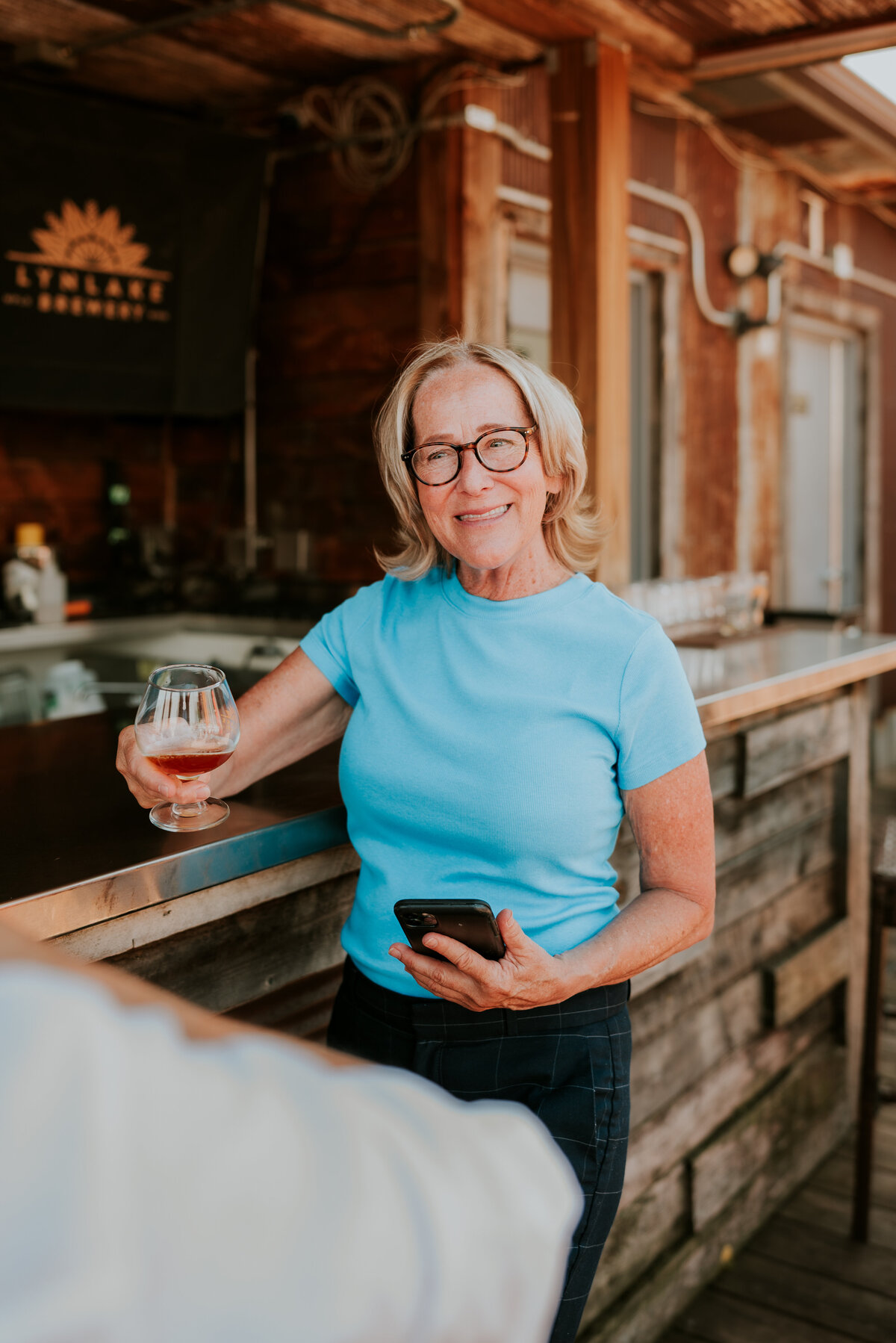 Relaxed and confident woman in turquoise shirt holding a wine glass at a warm, inviting bar setting.