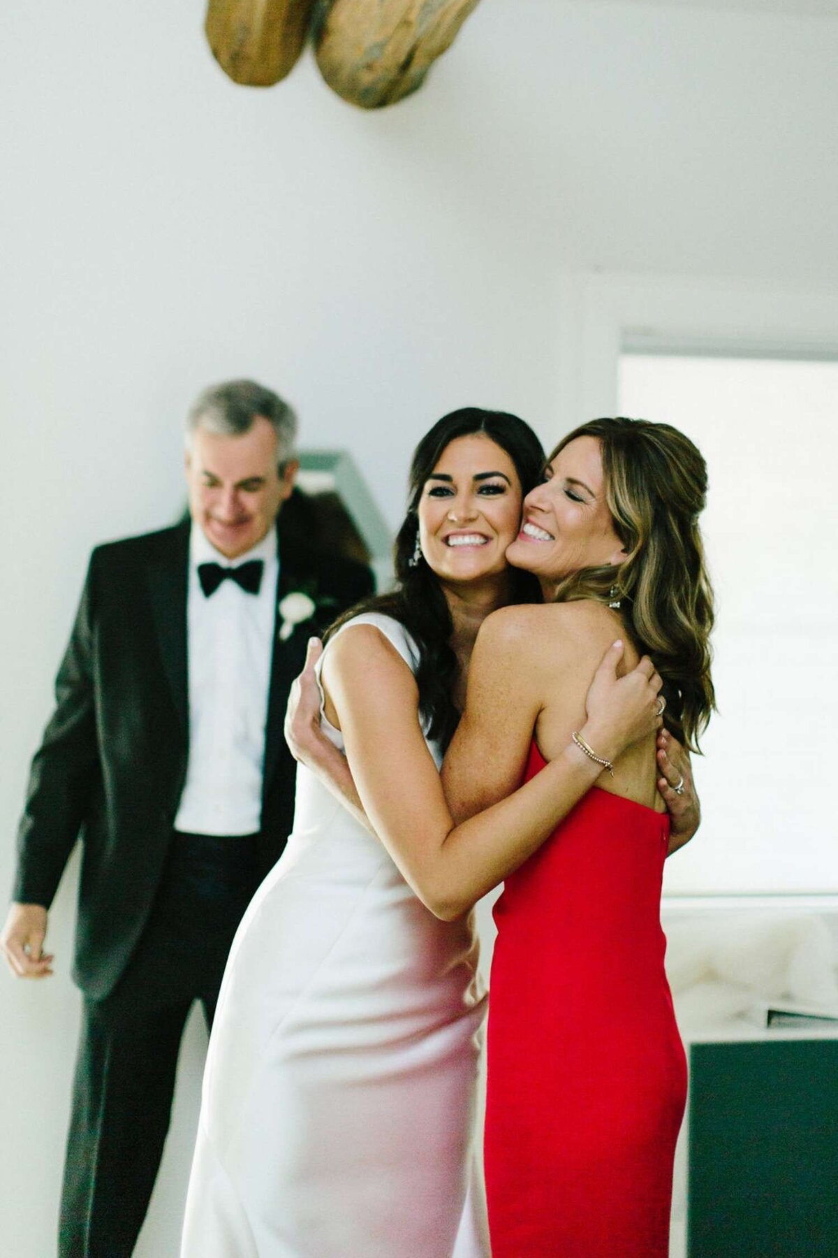Mother of the Bride in a Red Strapless Dresses hugs the Elegantly Dressed Bride before a Luxury Michigan Lakefront Golf Club Wedding.