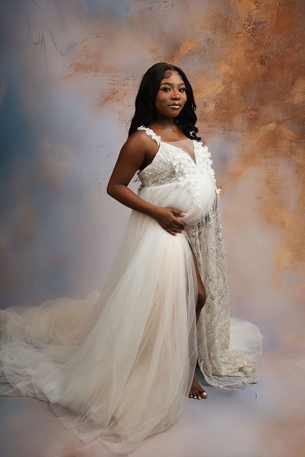 A mom to be stands in a photography studio holding her bump in a long white maternity gown