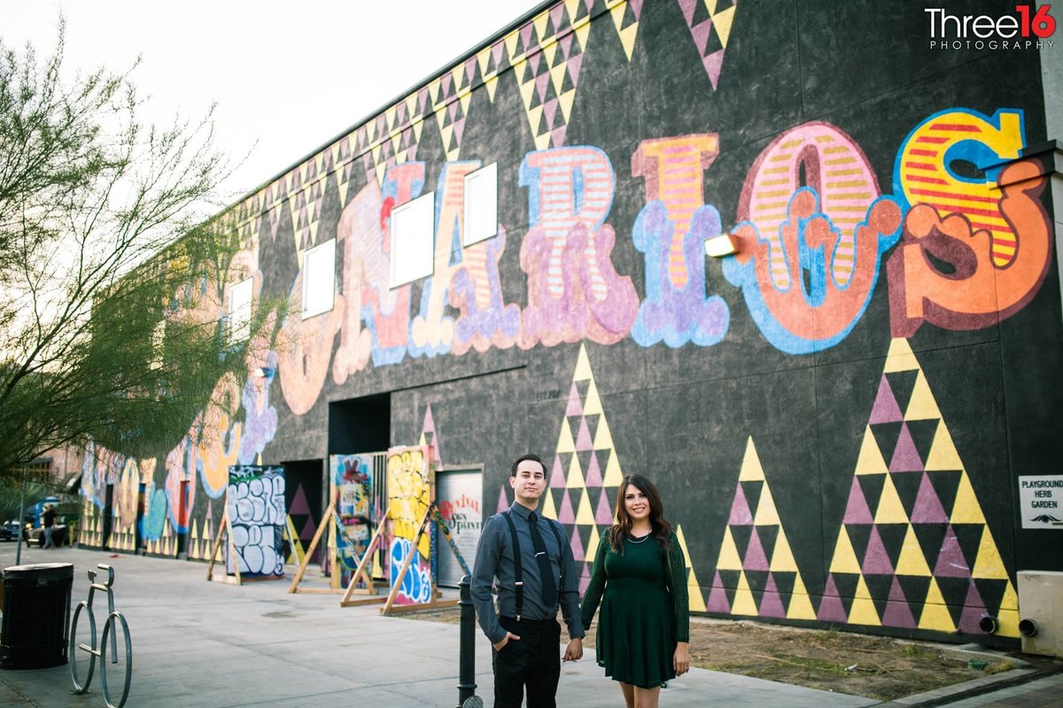 Engaged couple pose holding hands in front of a large wall mural in Santa Ana