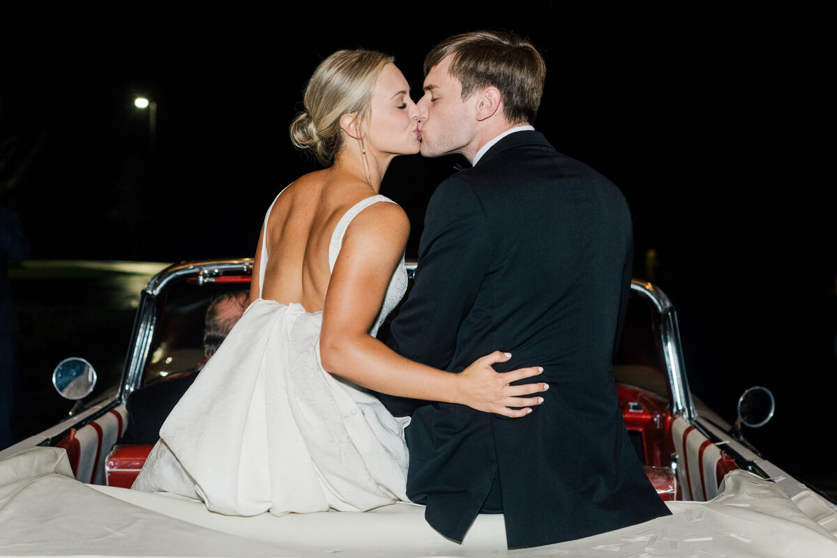 Bride and groom sitting on back of car kissing for wedding exit