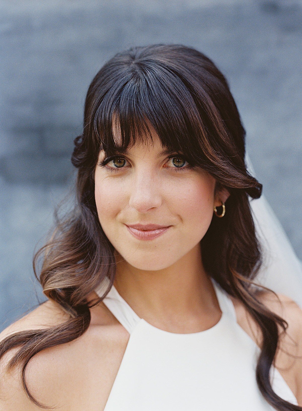 The bride, a brunette with long beachy waves smiles directly at the camera while she wears a veil and silk halter top wedding dress for her summer wedding at Clementine Hall in Nashville, TN