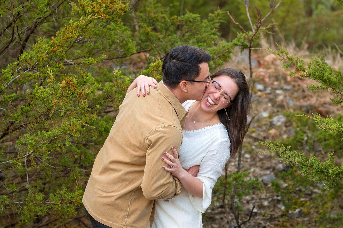 Bride and groom laughing near trees while groom kisses bride's cheek