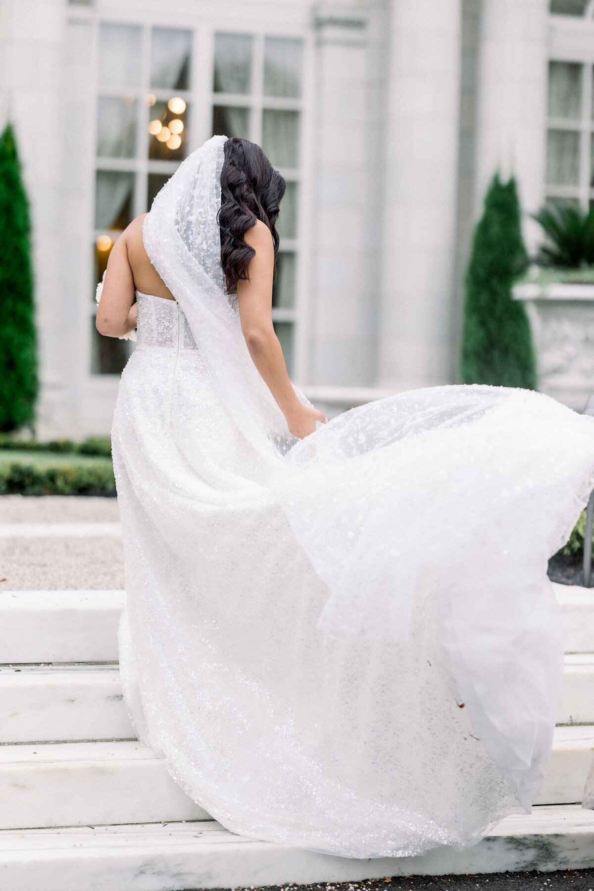 sophisticated bbride poses for glamorous bridal portrait  outside during wedding day portraits outside of rosecliff mansion in newport rhode island, the train of her gown gets swept by the wind