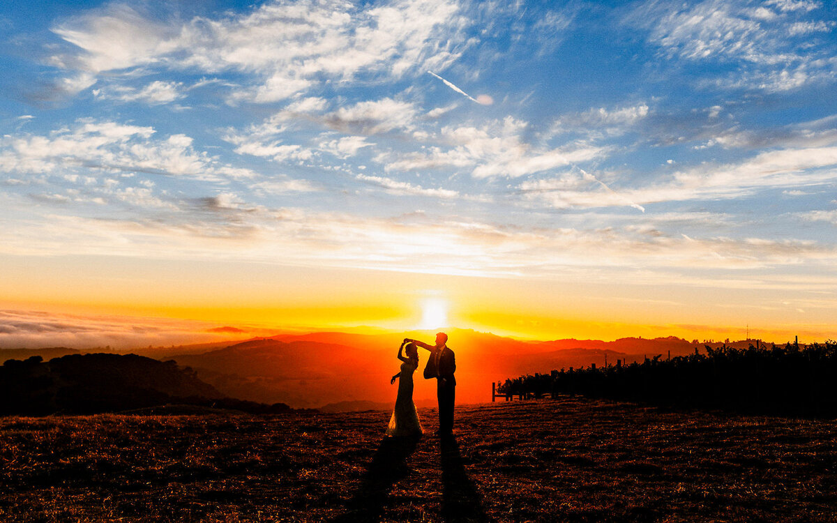 Bride and groom share a romantic dance at sunset on a mountaintop, silhouetted against a glowing orange sky and blue clouds.
