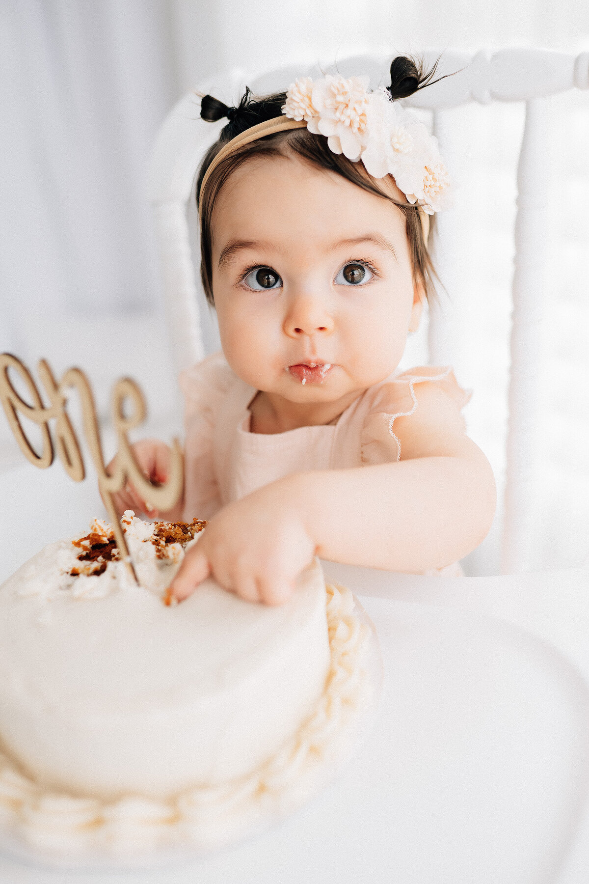 baby girl sitting in a white highchair with a cake for her first birthday milestone