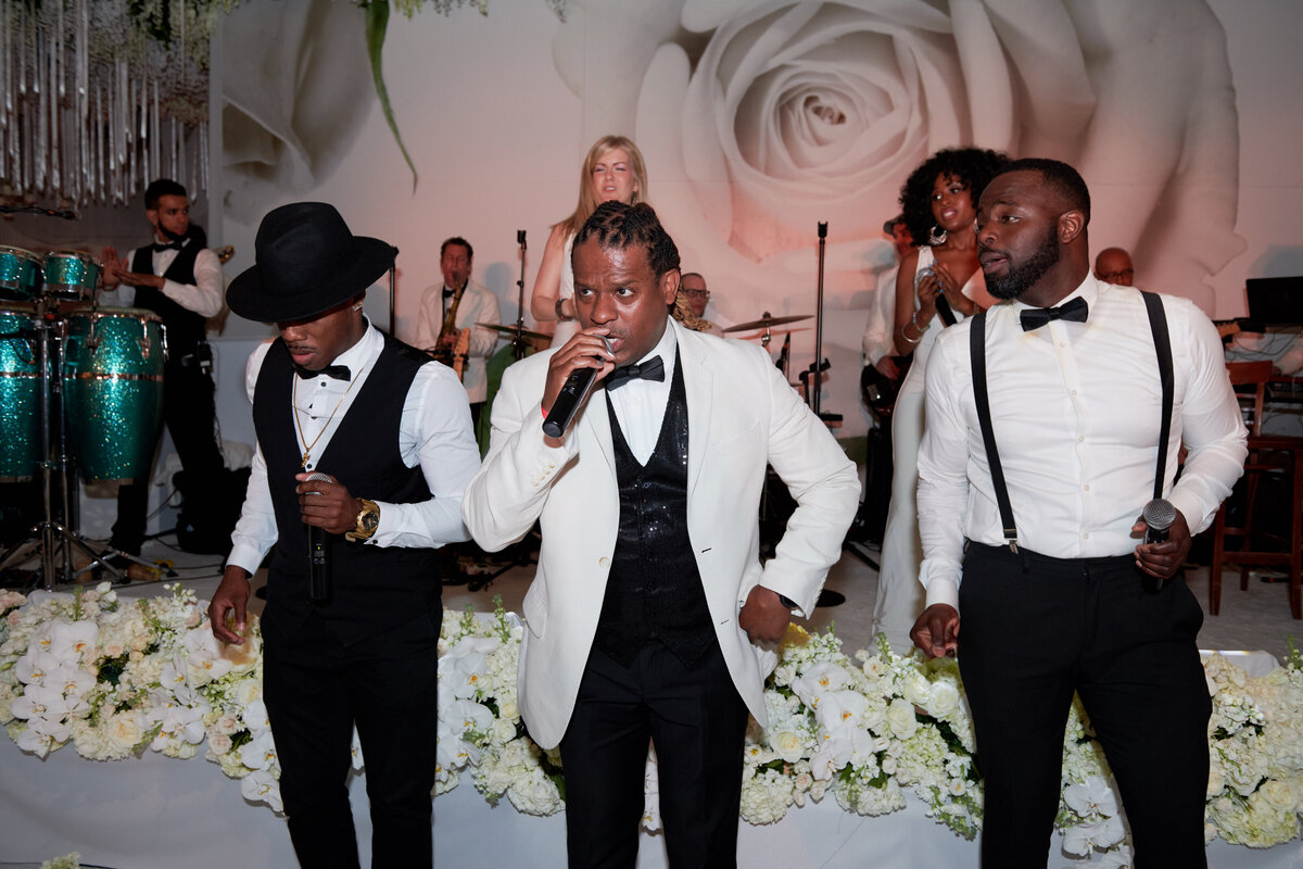 The photo was taken at the iconic New York Public Library in New York City by photographer Christian Oth. The band, consisting of three male singers dressed in black and white formal attire, can be seen performing on stage. In the background, a large white rose adds an elegant touch to the scene. Additionally, female musicians and other band members are visible playing various instruments, complemented by white flowers lining the stage. East West Films are the videographers capturing this memorable performance.