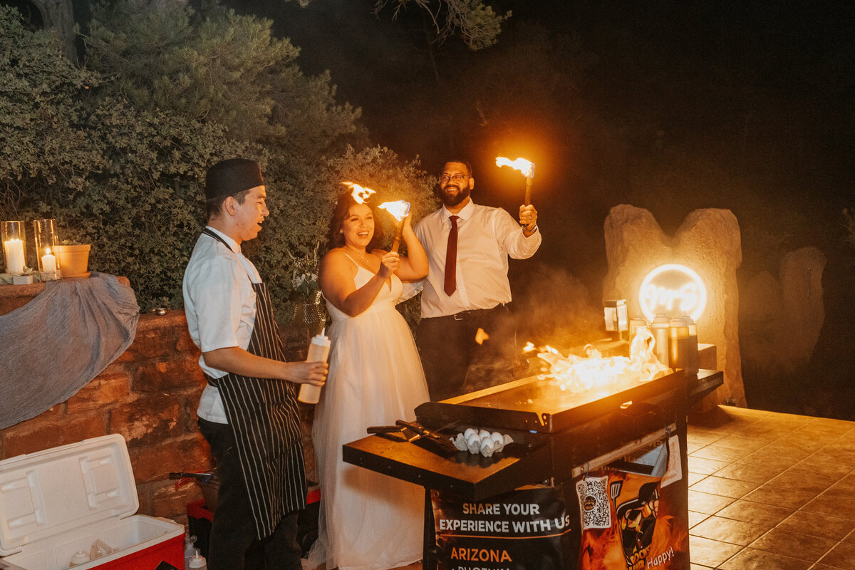 A newlywed couple standing in front of a hibachi grill holding flaming sticks 