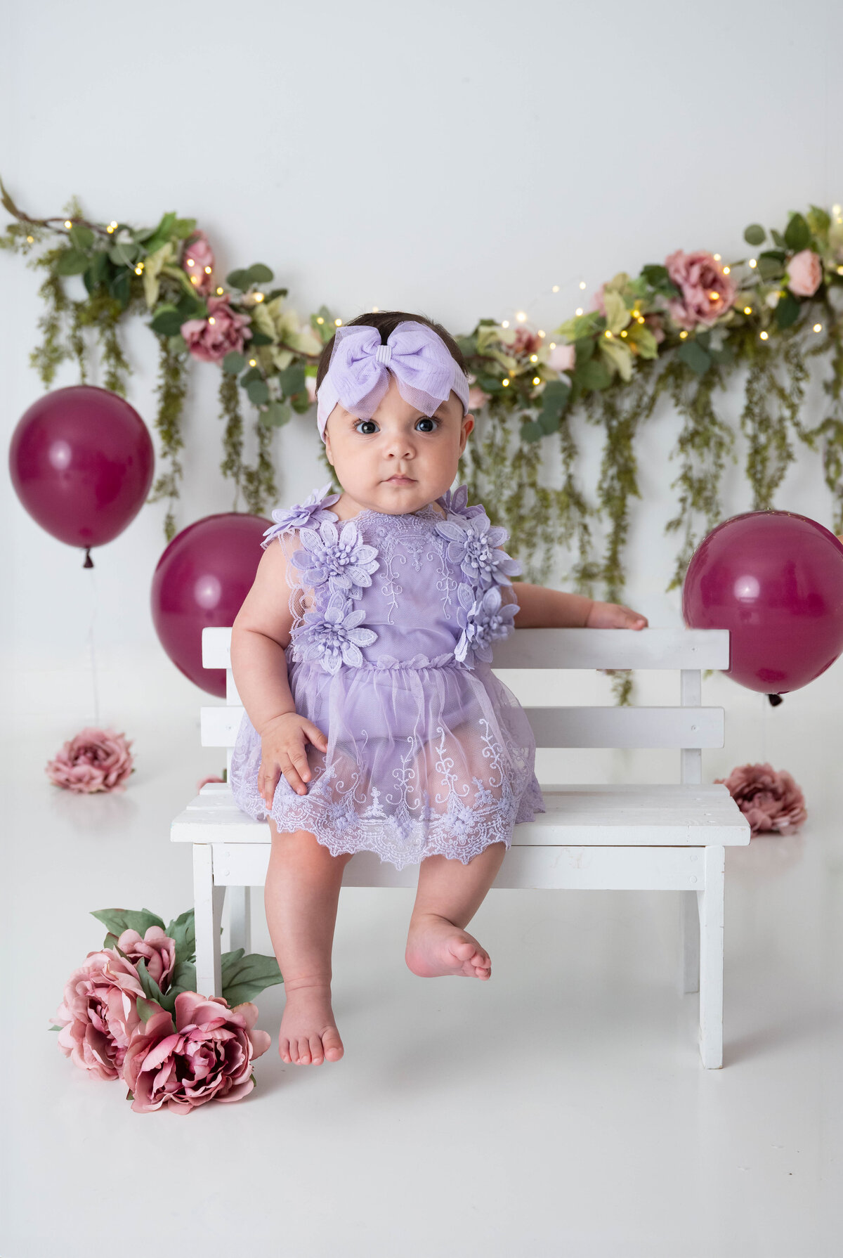 Baby girl in lilac lace dress and matching headband sitting on a white bench with mauve flowers and balloons in the background.