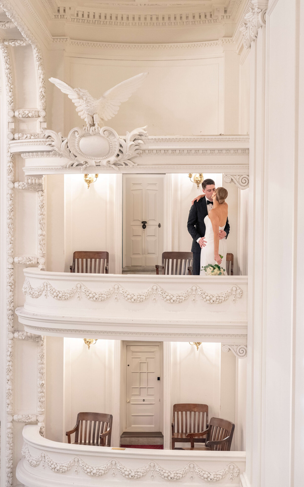 A bride and groom stand closely together on an ornate white balcony in an elegant venue with classical architectural details, including a carved eagle decoration above them. The interior features decorative moldings and wooden chairs.