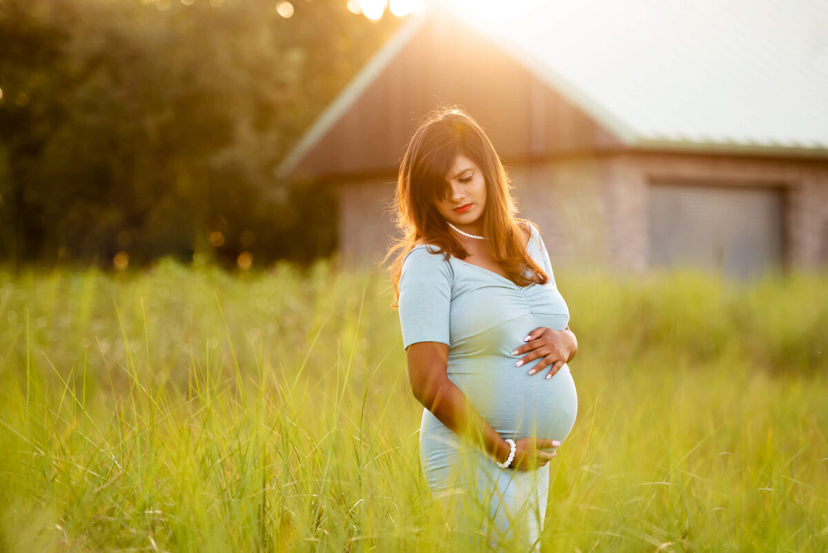 Woman standing in tall grass with the sun setting behind for her beach maternity photos.