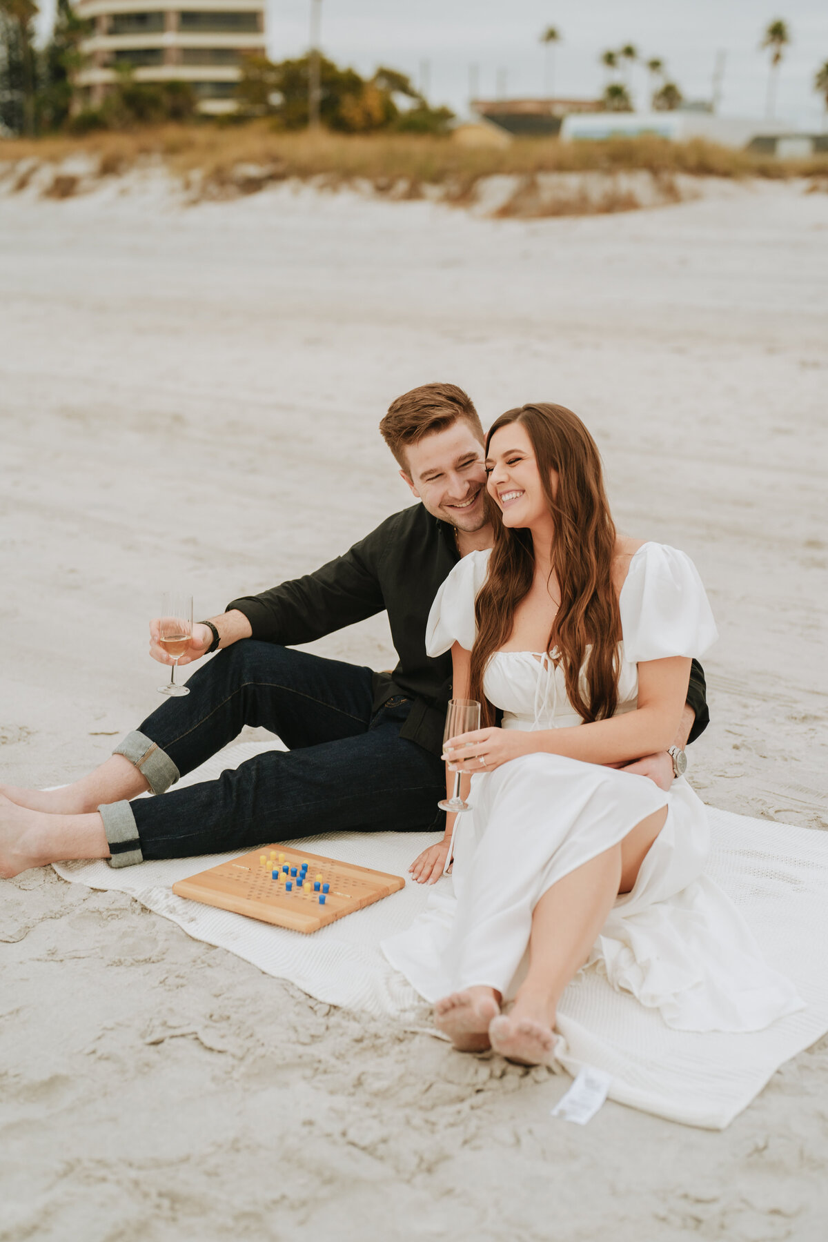 couple sitting on blanket at the beach during engagement session with champagne