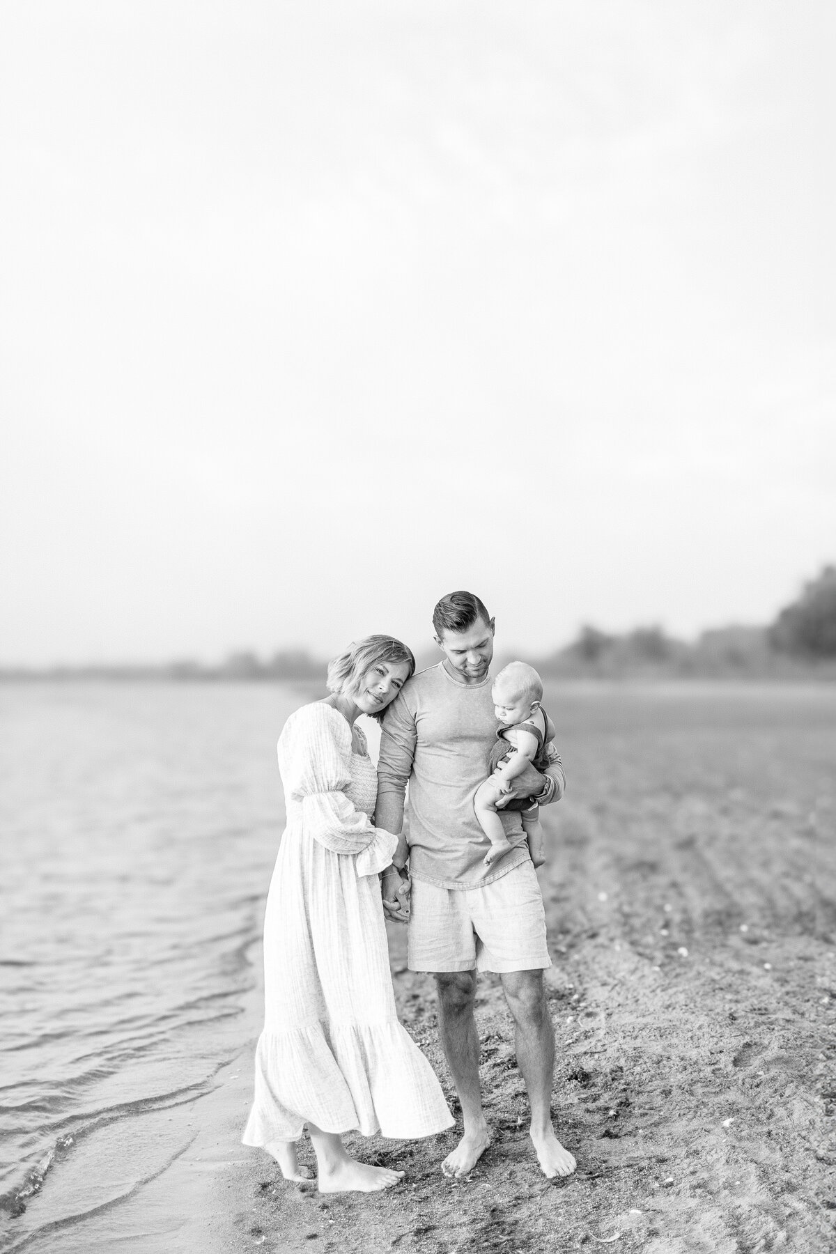 Black and white photo of parents standing on the beach with their baby boy during a 6-month milestone session at a Kansas City lake