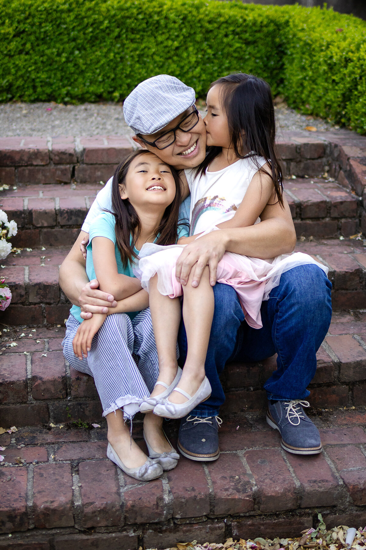 Golden hour portrait of father hugging  sisters two daughters girls hugging in rose garden – joyful, natural light family photography by Ellobelle Photography in the San Francisco Peninsula kohl mansion Burlingame