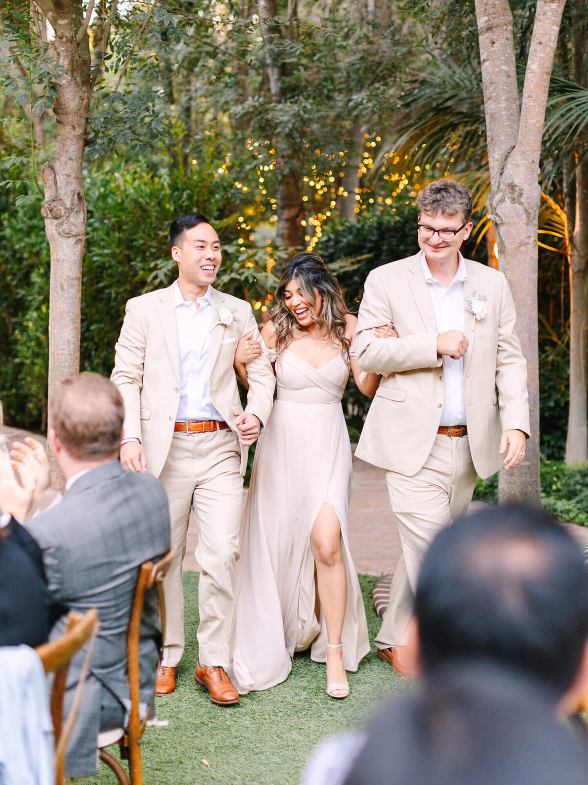 A bridesmaid in a beige dress walks arm-in-arm with two groomsmen in matching suits at a wedding reception.
