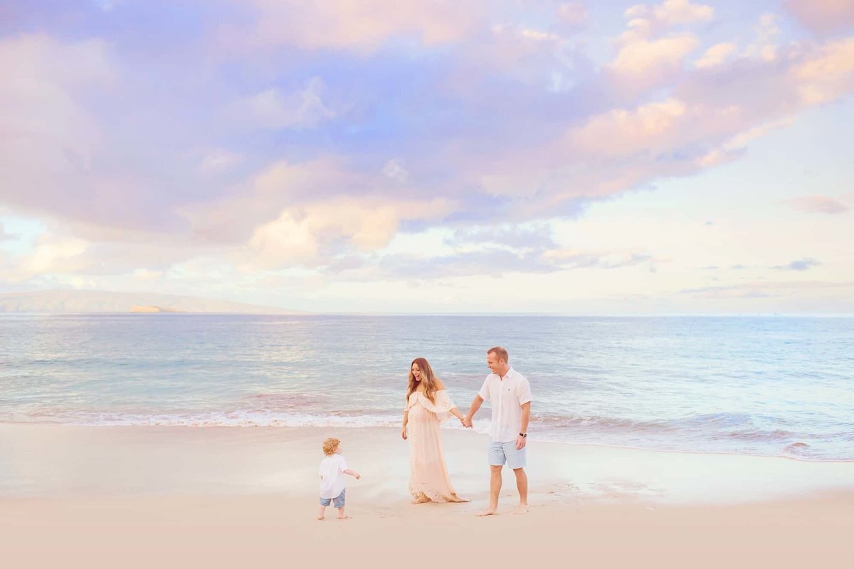 Family of three with pregnant mother in pink dress hold hands on the beach at sunrise in Wailea during a carefree family photo session