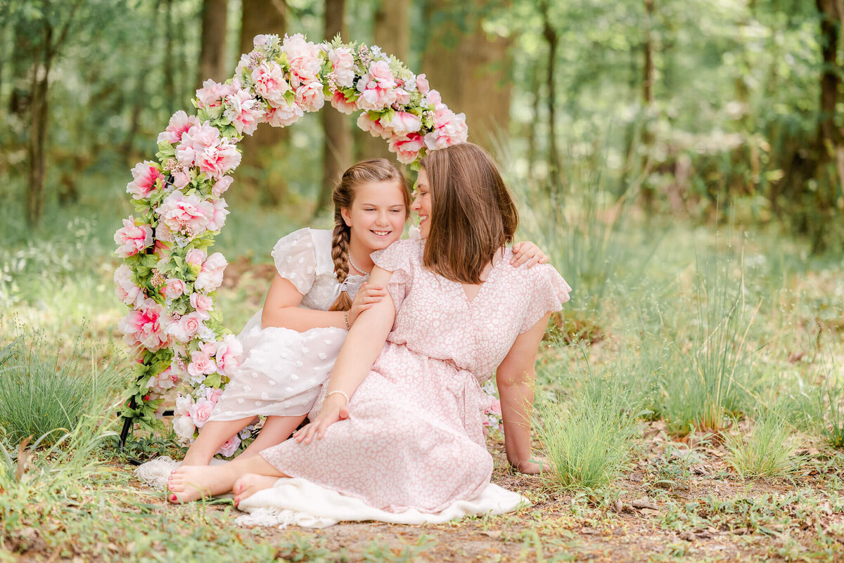 A mother and daughter wearing white and pink snuggle close during a Mother's Day photoshoot with a floral hoop. Photo by Justine Renee Photography.