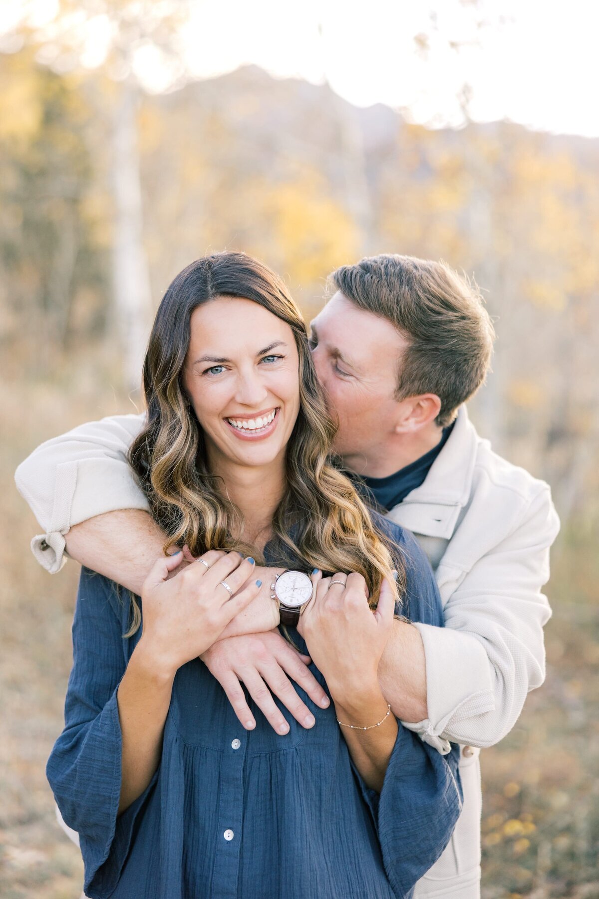 This is a closeup of a young boyfriend and girlfriend. His arms are wrapped around her chest. He is kissing her forehead while she smiles at the camera.