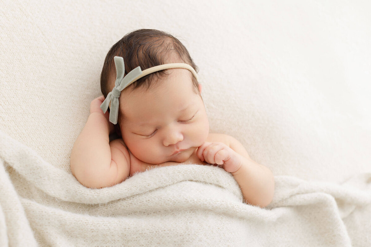 baby sleeping under a soft, cozy beige blanket with hands up by her head. She is wearing a sage green velvet bow