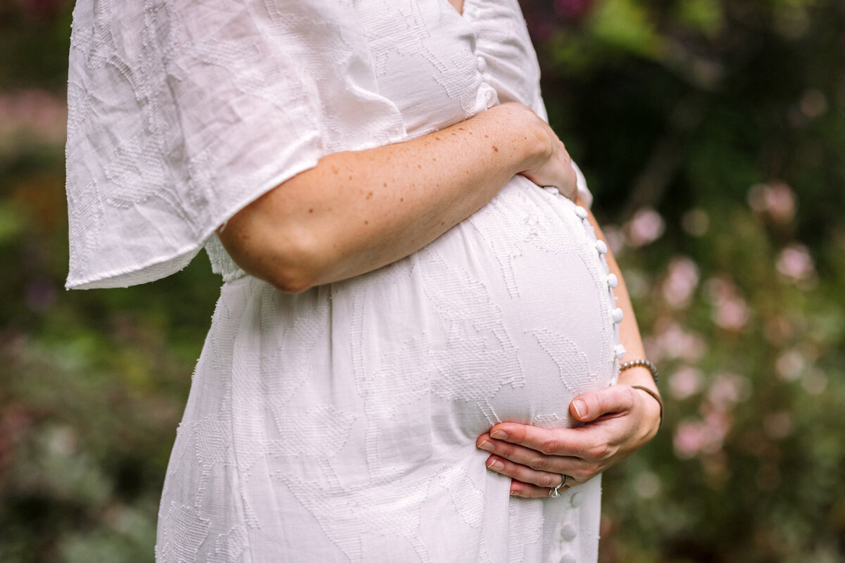 pregnant woman in white dress holding her baby bump in front of greenery with pink flowers in Oakland gardens
