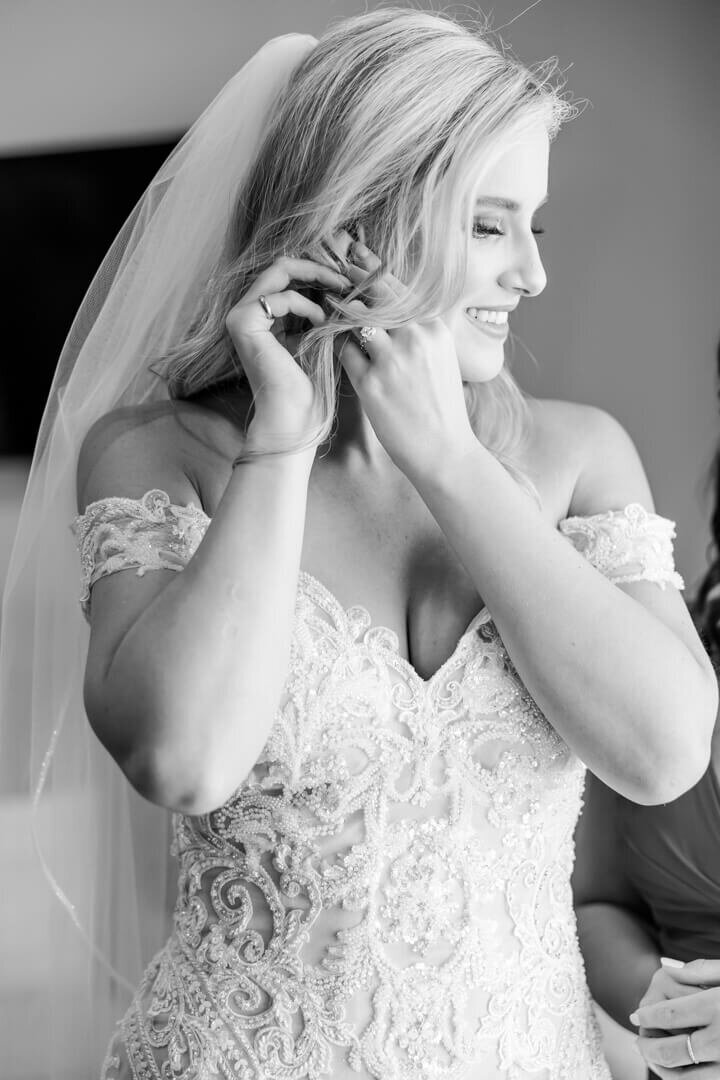 A black and white Arkansas wedding photography image of a bride putting on her earrings while smiling