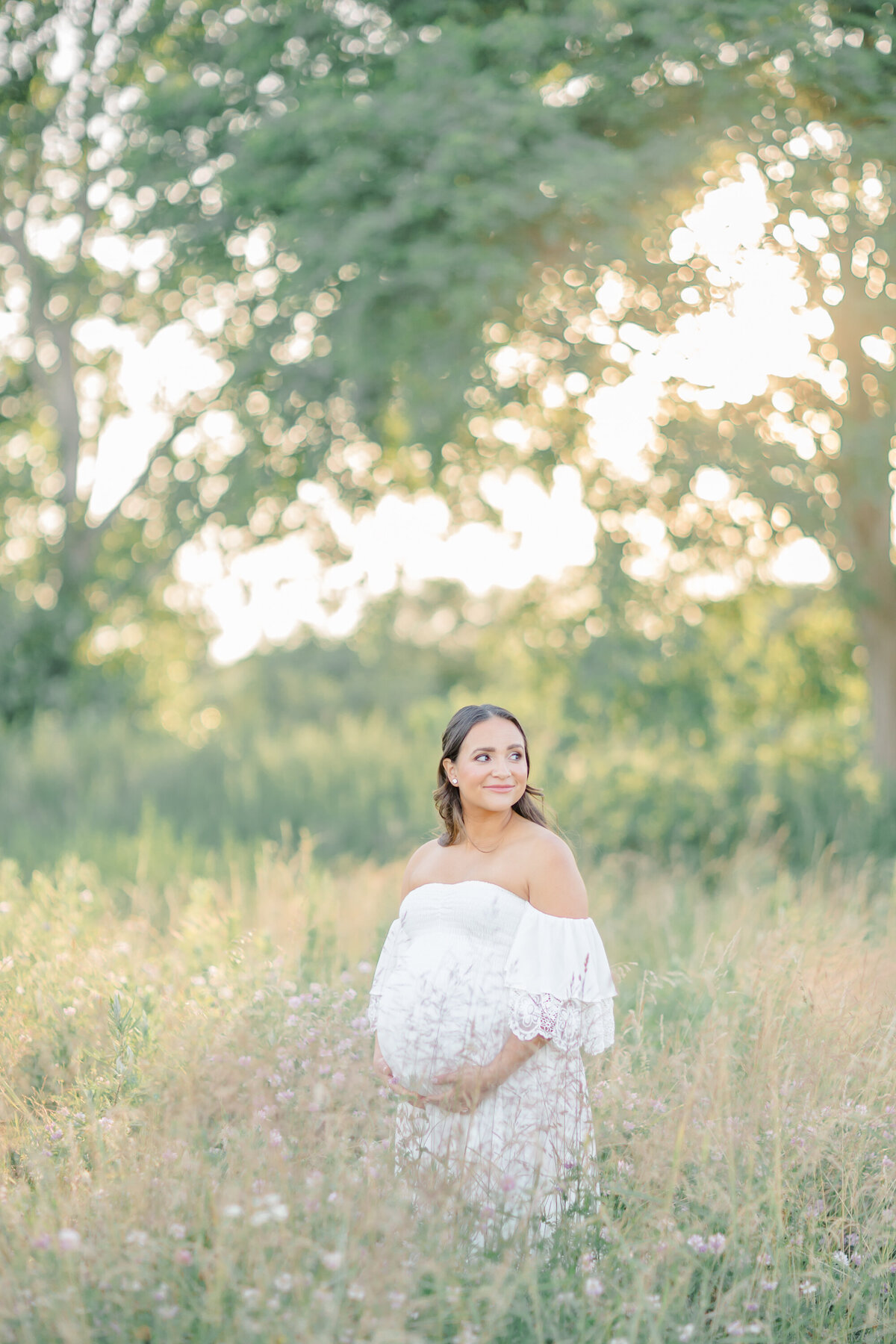 Expecting mother in a soft, flowing gown, standing in the middle of an open field with the sun shining brightly.