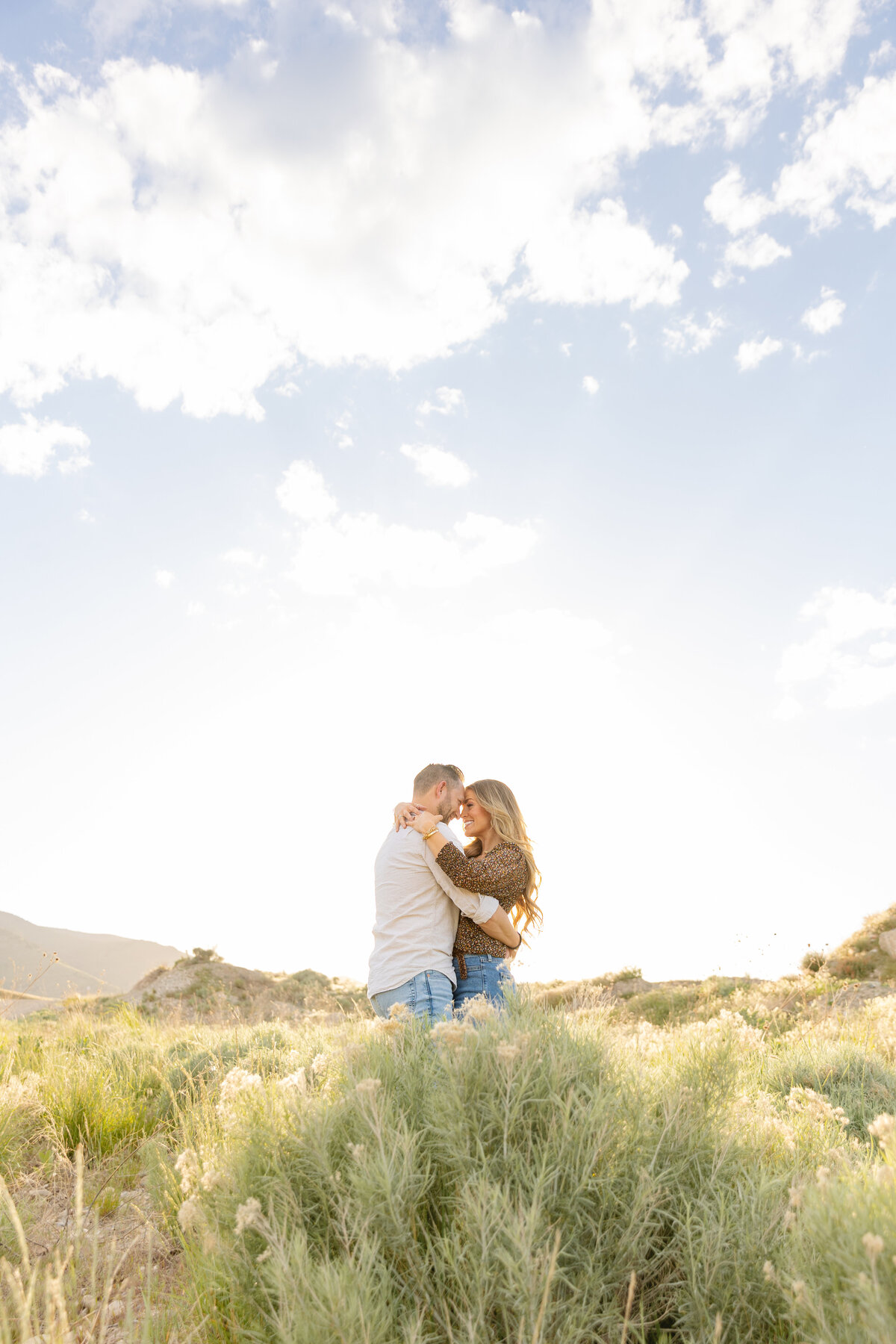 A couple stands together in a wide-open grassy landscape, embracing as the sun sets behind them. The warm glow of the evening light enhances the romance of the moment.
