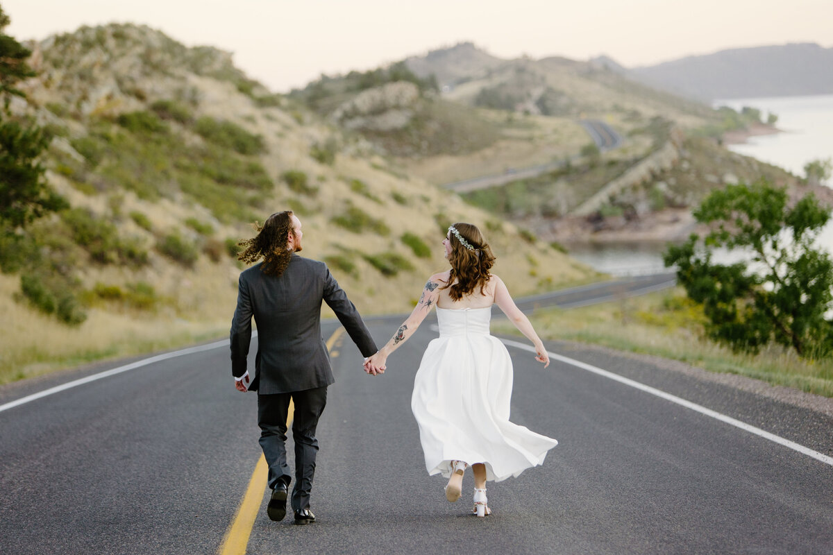 A joyful couple runs hand in hand down a winding road at Horsetooth Reservoir, soaking in the excitement of their elopement.